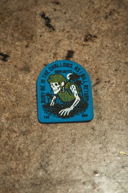 A rugged PVC patch by Born United Private Label shows a skeleton with seaweed and helmet, gripping a trident, plus the text: “Bury me in the shallows, but I’ll return. Born united. TRD. BNM.” Product: The Shallows PVC Patch.