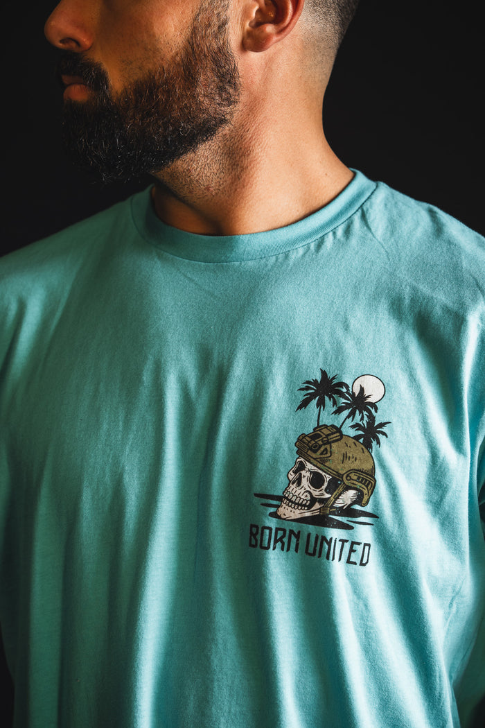 A bearded man wears a turquoise "The Shallows" t-shirt by Born United Private Label, featuring a skull in a helmet, palm trees, moon graphic, and "BORN UNITED" text on the chest.