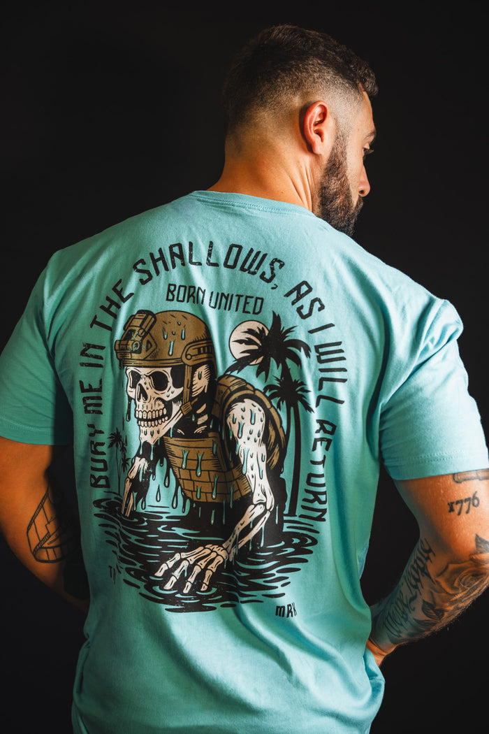 A tattooed man, seen from behind, wears Born United Private Label's "The Shallows" tee. The turquoise shirt features a skeleton in armor rising from water with palm trees and the text: "Bury me in the shallows, as I will return.